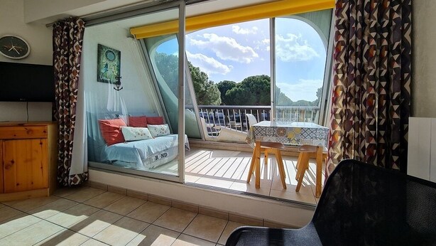 Une pièce dotée d’une grande porte-fenêtre coulissante donnant sur une terrasse extérieure. On y trouve un lit avec des oreillers et une petite table avec des chaises. Un téléviseur est fixé au mur.