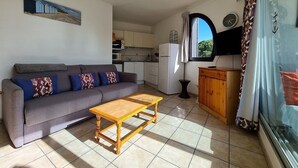 Un espace de vie compact comprenant un canapé, une table basse en bois, un coin cuisine avec des placards et des appareils électroménagers, et une fenêtre avec des rideaux.