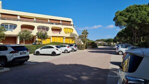 Un parking avec plusieurs voitures, un bâtiment à la façade jaune et beige, et des arbres en arrière-plan.