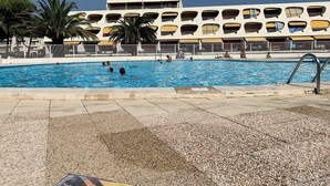 Une piscine où des personnes nagent et se détendent, entourée d’un bâtiment doté de nombreux balcons et d’un palmier.
