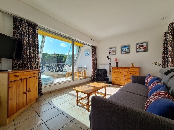 A living room with a sofa, wooden cabinet, and a television. There is a balcony with a table and chairs.