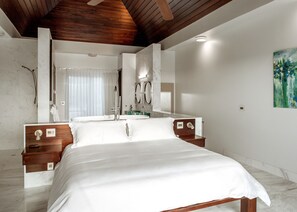 Quarto moderno com cama grande, cabeceira de madeira, closet espaçoso e banheiro com chuveiro e espelho amplo.