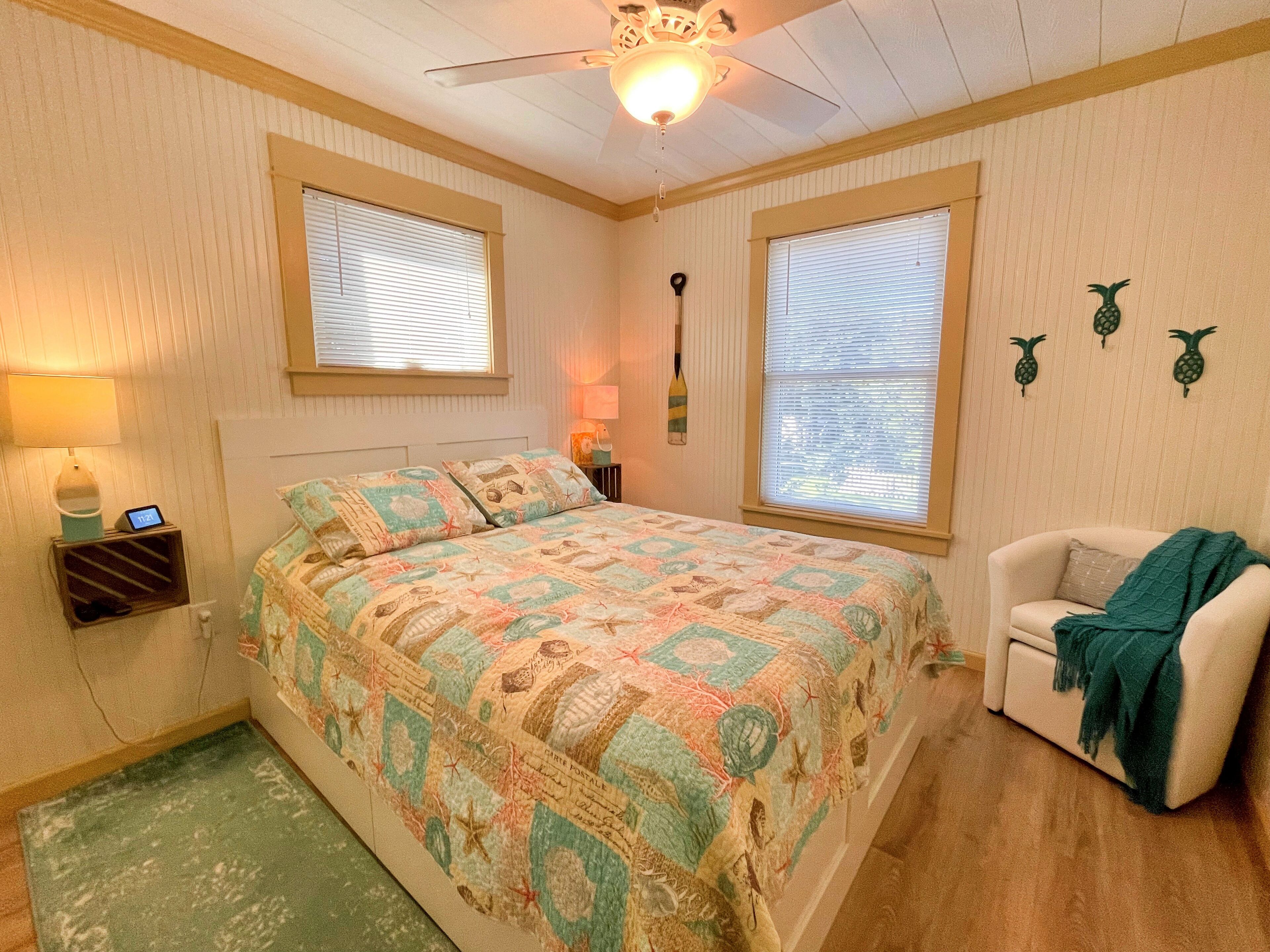 A bedroom with a bed, two windows with blinds, a ceiling fan, a nightstand with a lamp, a wall clock, and a small shelf with a bottle and a cup.