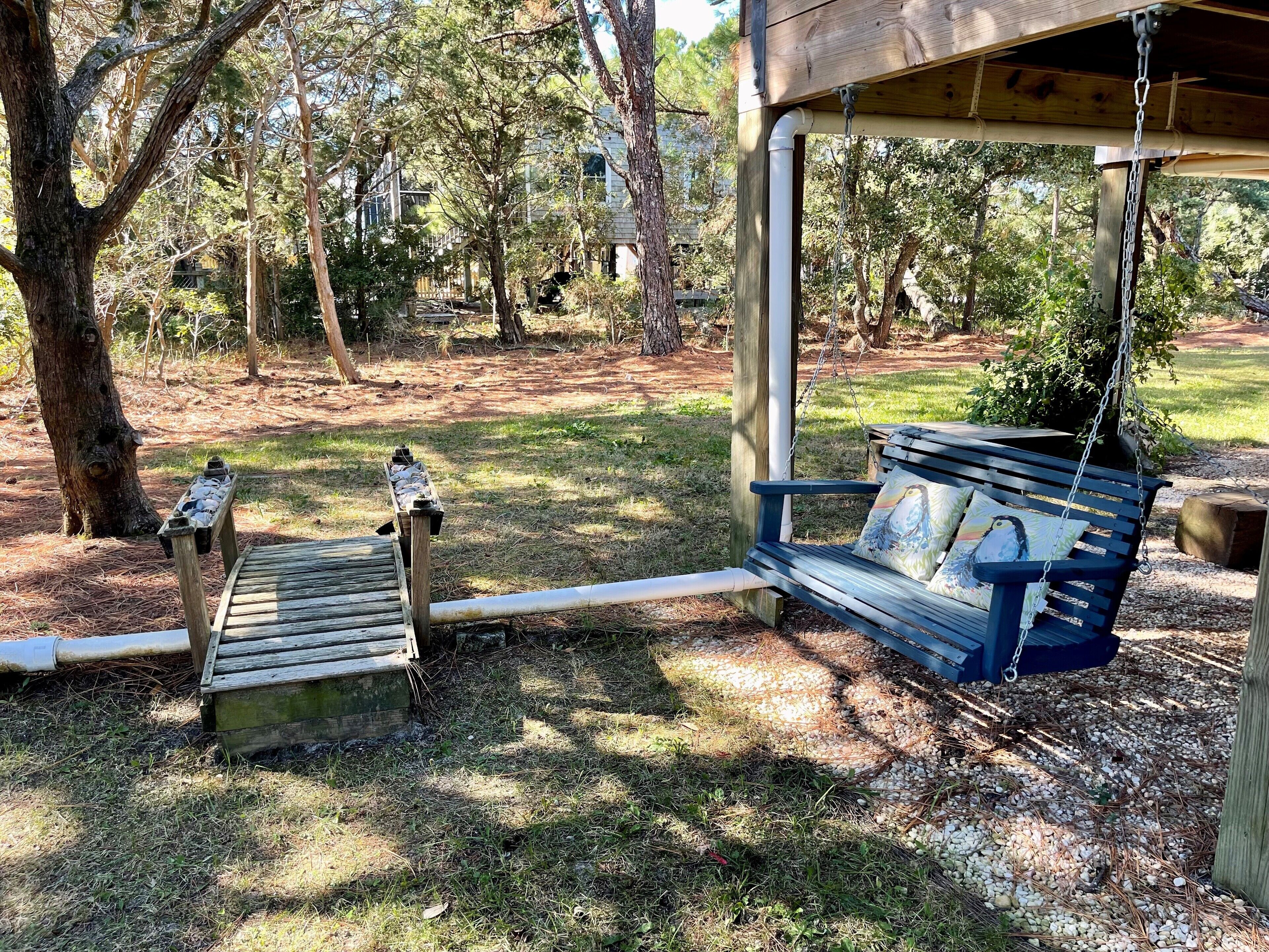 A wooden bench with a swing seat, a small wooden table, and a metal bench rail in a wooded outdoor area.