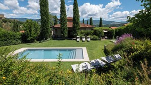 Una piscina con sillones de descanso, rodeada de vegetación y árboles altos, con una casa al fondo.