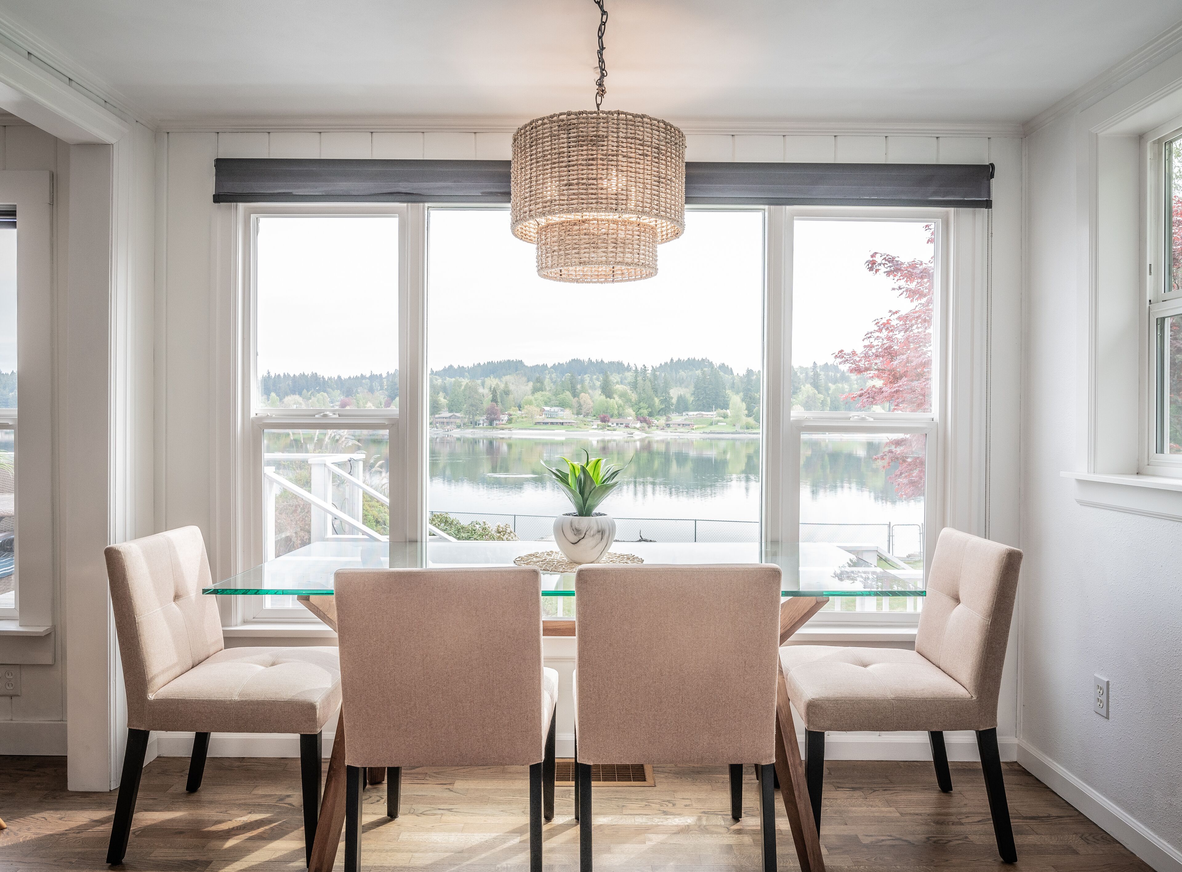 Un comedor con una mesa y sillas, ventanas amplias con vista a un lago y árboles, y una lámpara colgante.