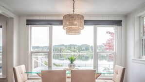 Un espace repas comprenant une table et des chaises, de grandes fenêtres donnant sur un lac et des arbres, ainsi qu’un luminaire suspendu.