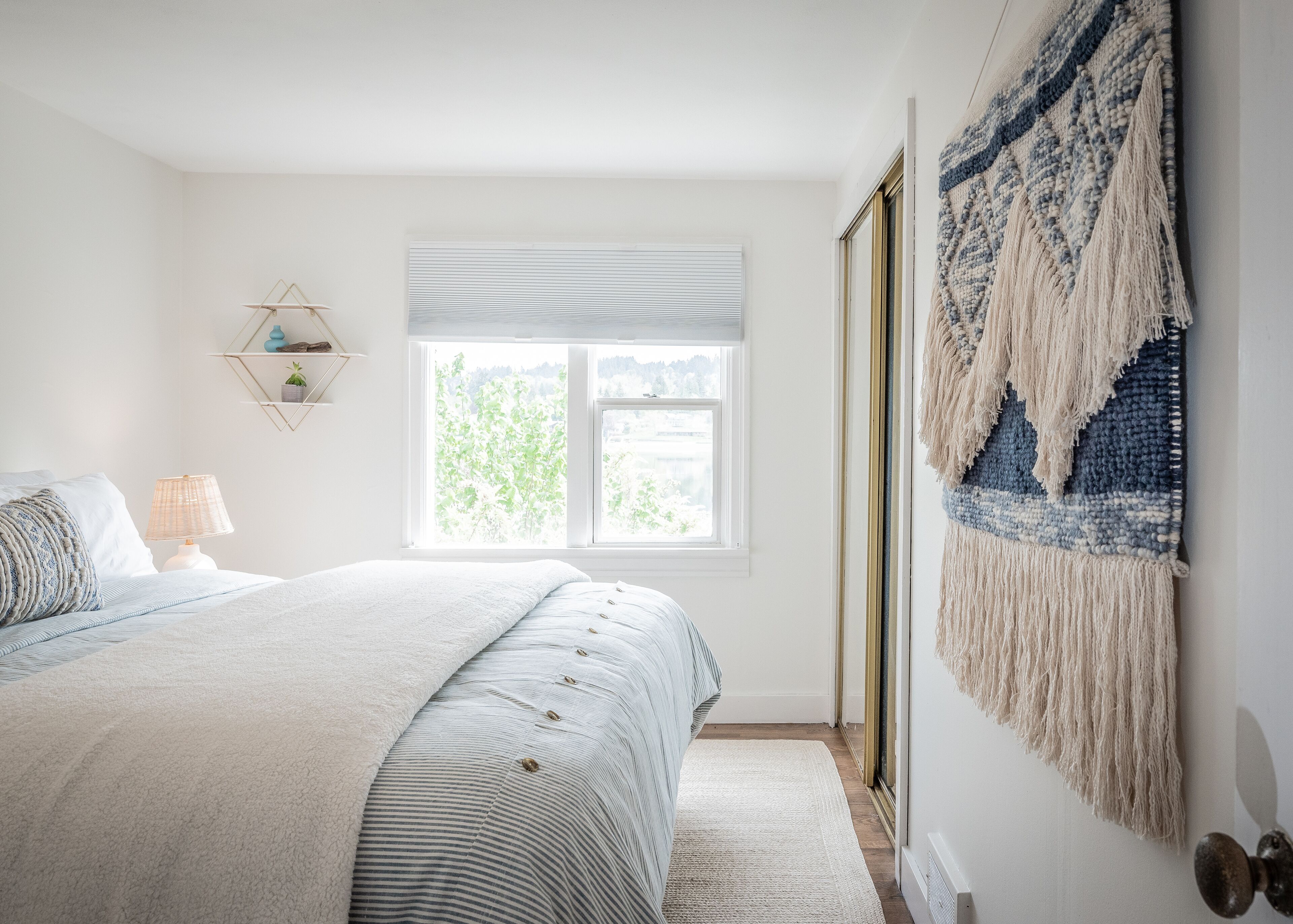 A bedroom with a large bed, a window with blinds, a wall-mounted macrame piece, and a small shelf with decorative items.