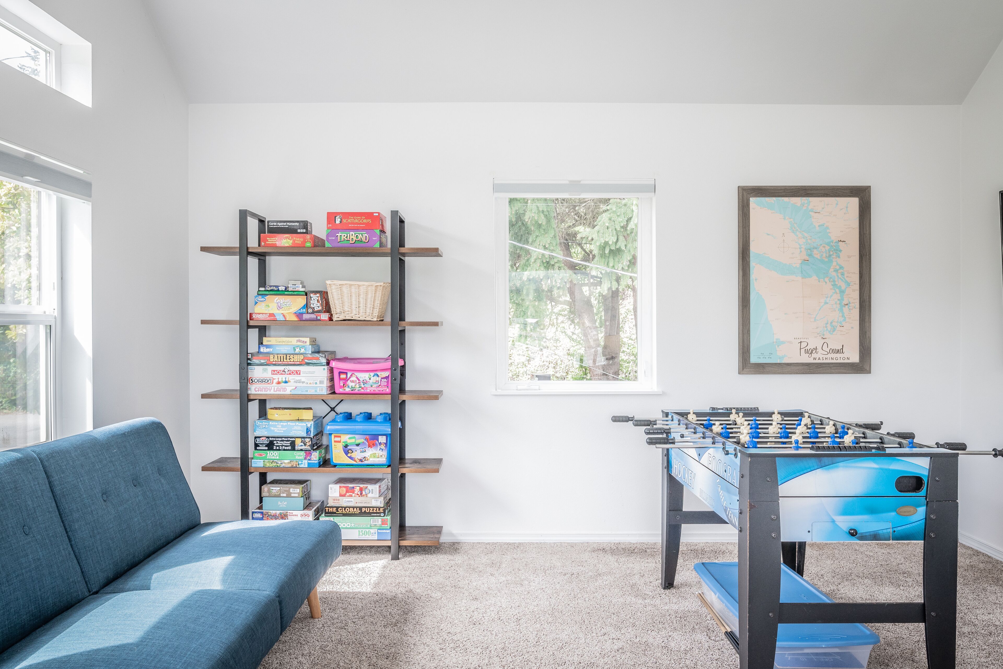 A room with a sofa, a bookshelf with games, a window with a view of trees, and a foosball table.