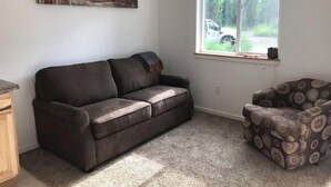 Ein Wohnzimmer mit einem dunkelbraunen Ledersofa, einem gemusterten Sessel, einem Gemälde an der Wand und einem Fenster mit Blick auf ein Fahrzeug und Grünflächen.