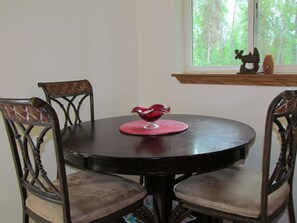 A round wooden dining table with a red decorative bowl in the center, surrounded by four upholstered chairs. A window with a view of trees is visible in the background.