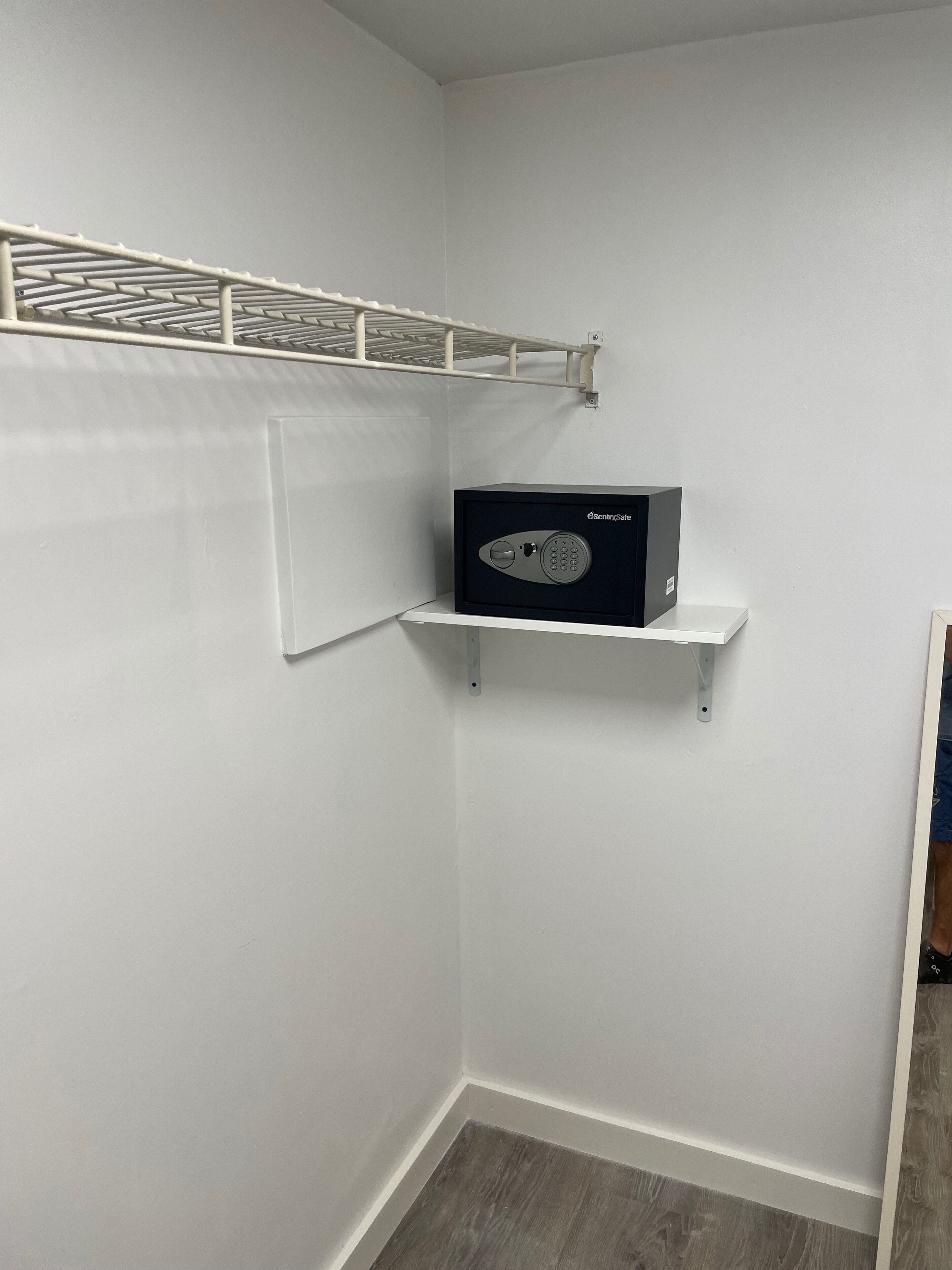 A corner closet with a metal shelf holding a black safe, a white wall, and a wooden floor.