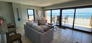A living room with a gray sofa, wooden chairs, a dining table, and a view of the beach through large sliding glass doors.