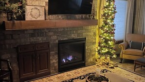Un salon doté d’une cheminée en pierre, d’un téléviseur à écran plat fixé au-dessus, d’un sapin de Noël et d’un coin salon confortable agrémenté d’un tapis à motifs.