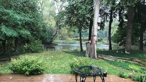 Un patio in legno con tavolo e sedie in metallo, circondato da vegetazione rigogliosa e un fiume sullo sfondo.