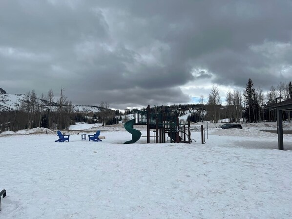 Zona exterior nevada con un parque infantil, que incluye un tobogán y columpios, y un coche aparcado cerca.