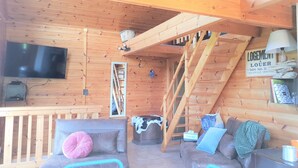 A wooden cabin interior with a TV mounted on the wall, a staircase, a couch with cushions, and a sign indicating the room is a living area. - Wooden chalet with magnificent view (Neuviller-la-roche)
