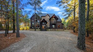 Une grande cabane rustique en rondins de bois, avec des fondations en pierre, entourée d’une allée de gravier et de grands arbres.