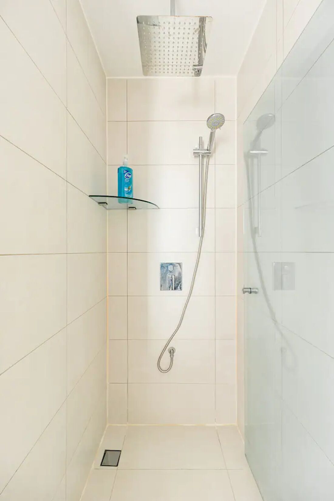 Un pequeño box de ducha con azulejos, un cabezal de ducha manual y un grifo empotrado en la pared. Hay un estante de cristal con una botella de champú y un pequeño marco de fotos.