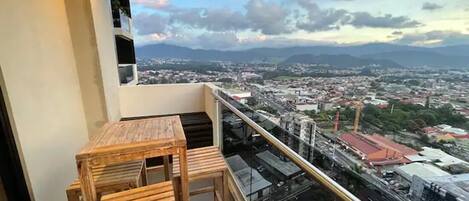 A balcony with a wooden table and chairs, overlooking a cityscape with mountains in the distance.