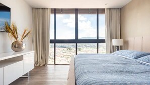 A modern hotel room with a large window, a bed, a bedside table, and a lamp.