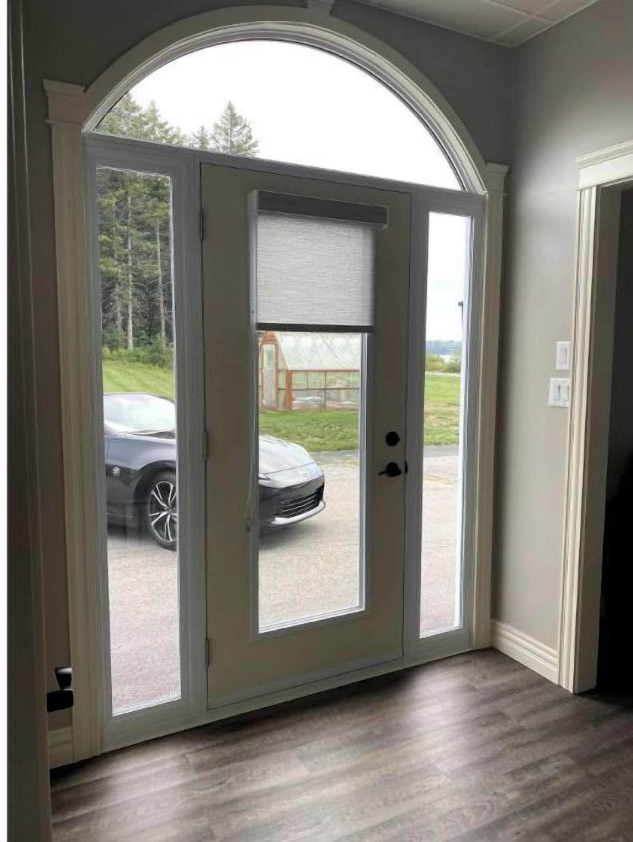 A door with a view of a car outside, a window with a blind, and a wooden floor.