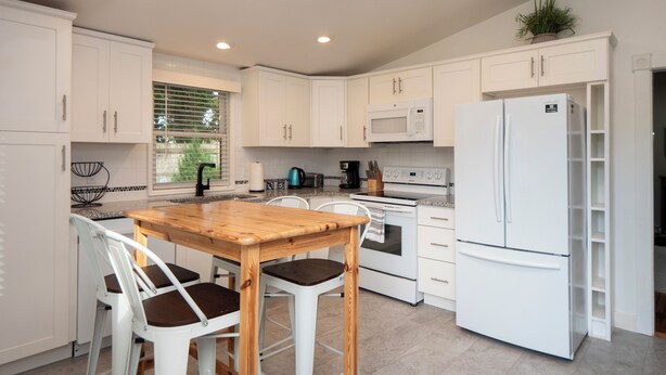A modern kitchen with white cabinets, a wooden table with chairs, a white refrigerator, a microwave, and a stove. - Cheerful two bedroom cottage with Parisian patio! (Middletown)