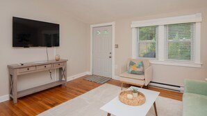 A living room with a flat-screen TV mounted on the wall, a wooden console table, a beige sofa with a patterned cushion, a white coffee table with decorative items, and a window with blinds. - Cheerful two bedroom cottage with Parisian patio! (Middletown)
