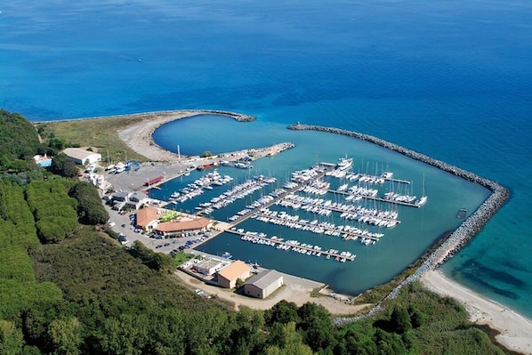 An aerial view of a marina with numerous boats docked, surrounded by greenery and a clear blue sea.