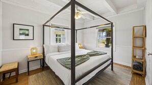 Een hemelbed met een plafondventilator, een nachtkastje met een lamp, een raam met gordijnen en een houten plank.
