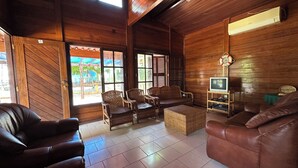 A wooden interior with leather sofas, wicker chairs, a television on a stand, and a view through large windows. - Suíte, Vila Alice, Luanda - Angola (Luanda)