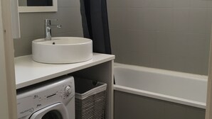 Een kleine badkamer met een witte wastafel, een ingebouwde wasmachine, een droogrek en een bad.