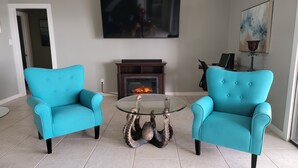 Een woonkamer met twee turquoise fauteuils, een glazen salontafel met een decoratief beeld, een open haard, een flatscreen televisie en een schilderij aan de muur.