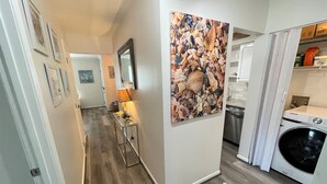 A hallway with a wooden floor, a painting of seashells on the wall, a lamp, and a Samsung washing machine.