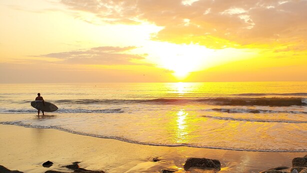 A beach scene with a surfer walking towards the ocean, waves, and a sunset.