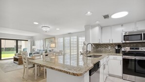 A modern kitchen with white cabinets, a granite countertop, stainless steel appliances, and a dining area with a large window. - Oceanfront | Ground Floor | Updated Amelia Surf and Racquet Condo (Fernandina Beach)