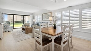 A spacious dining area with a wooden table and white chairs, large windows with blinds, and a chandelier. - Oceanfront | Ground Floor | Updated Amelia Surf and Racquet Condo (Fernandina Beach)