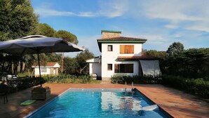 Una piscina rettangolare con acqua cristallina, circondata da un solarium piastrellato. Si vede un edificio a due piani con tetto in tegole e un piccolo balcone sullo sfondo.