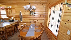 A wooden cabin interior with a dining table, chairs, a chandelier, and a window with blinds. - Grand Lake Cabin Retreat - Lake, Dock, & Woods (Vinita)