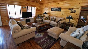 A cozy cabin-style living room with wooden walls and floors, a large window, a flat-screen TV, and a sign reading "Welcome to our Lake House." - Grand Lake Cabin Retreat - Lake, Dock, & Woods (Vinita)