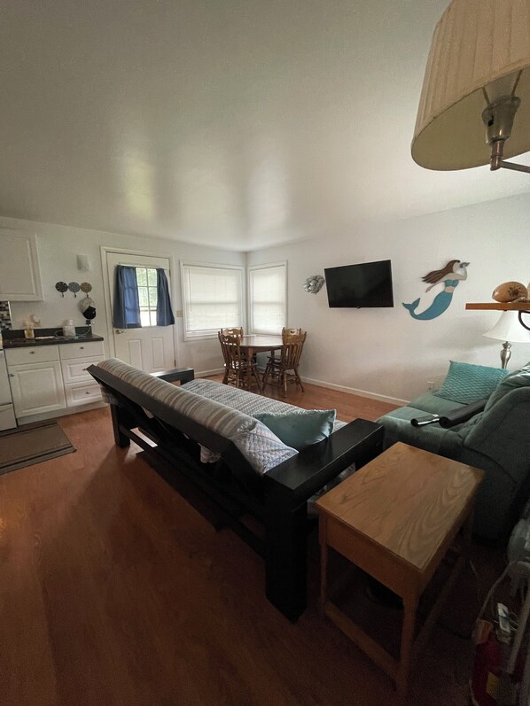 Ein kompakter Wohnraum mit Bett, kleinem Tisch, Sofa, Esstisch mit Stühlen, Fernseher und Fenster mit Jalousien.