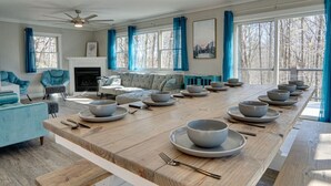 Amplia sala de estar con una larga mesa de comedor lista para una comida, una chimenea y ventanas grandes con cortinas.