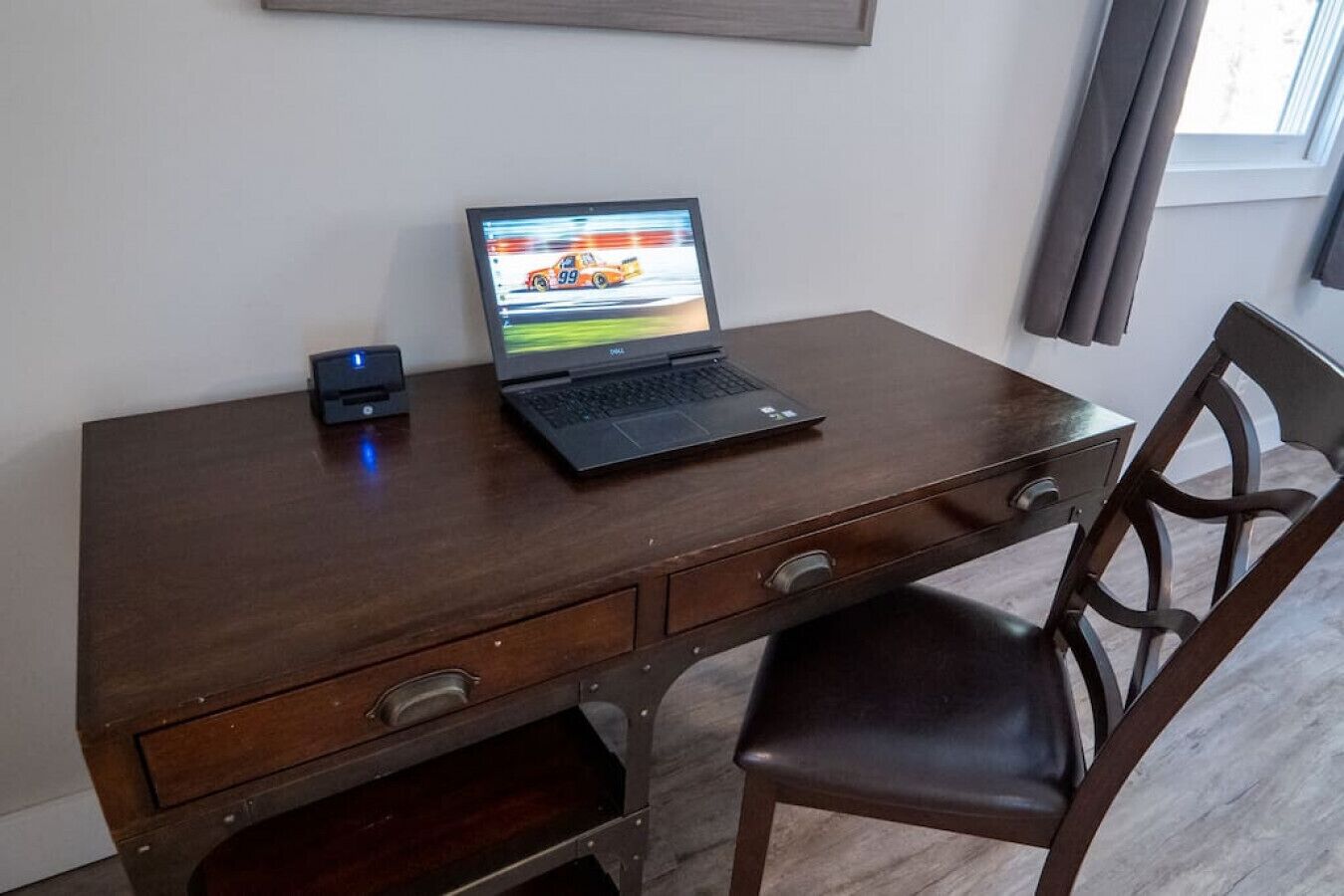 Un bureau en bois avec un ordinateur portable et un petit appareil, une chaise et une fenêtre avec des rideaux.