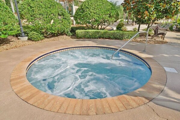A hot tub surrounded by a tiled edge and a stone border, set in a landscaped area with trees and a poolside lounge.