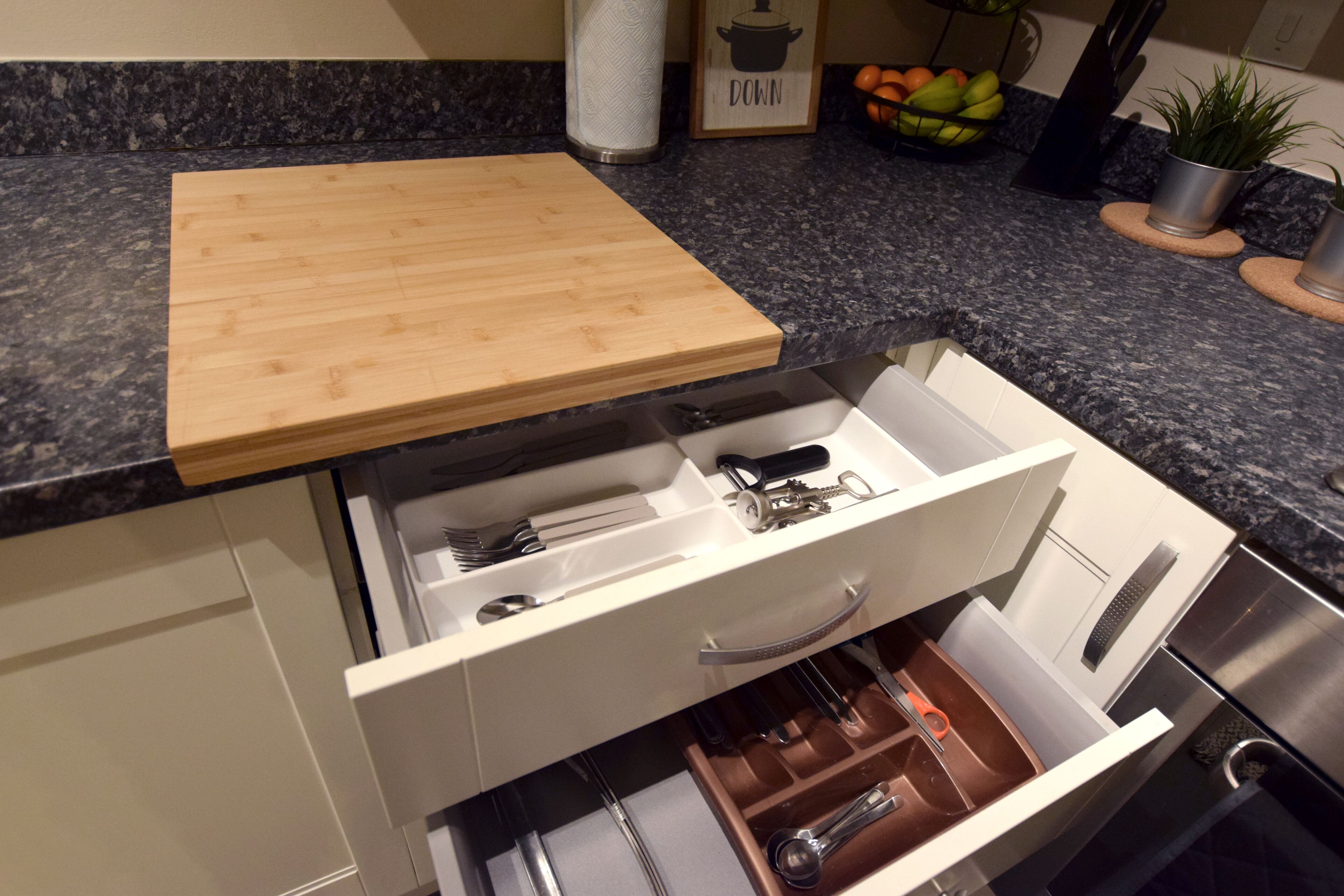 Una cocina con una tabla de cortar de madera en la encimera, un cajón blanco con utensilios y un frutero al fondo.