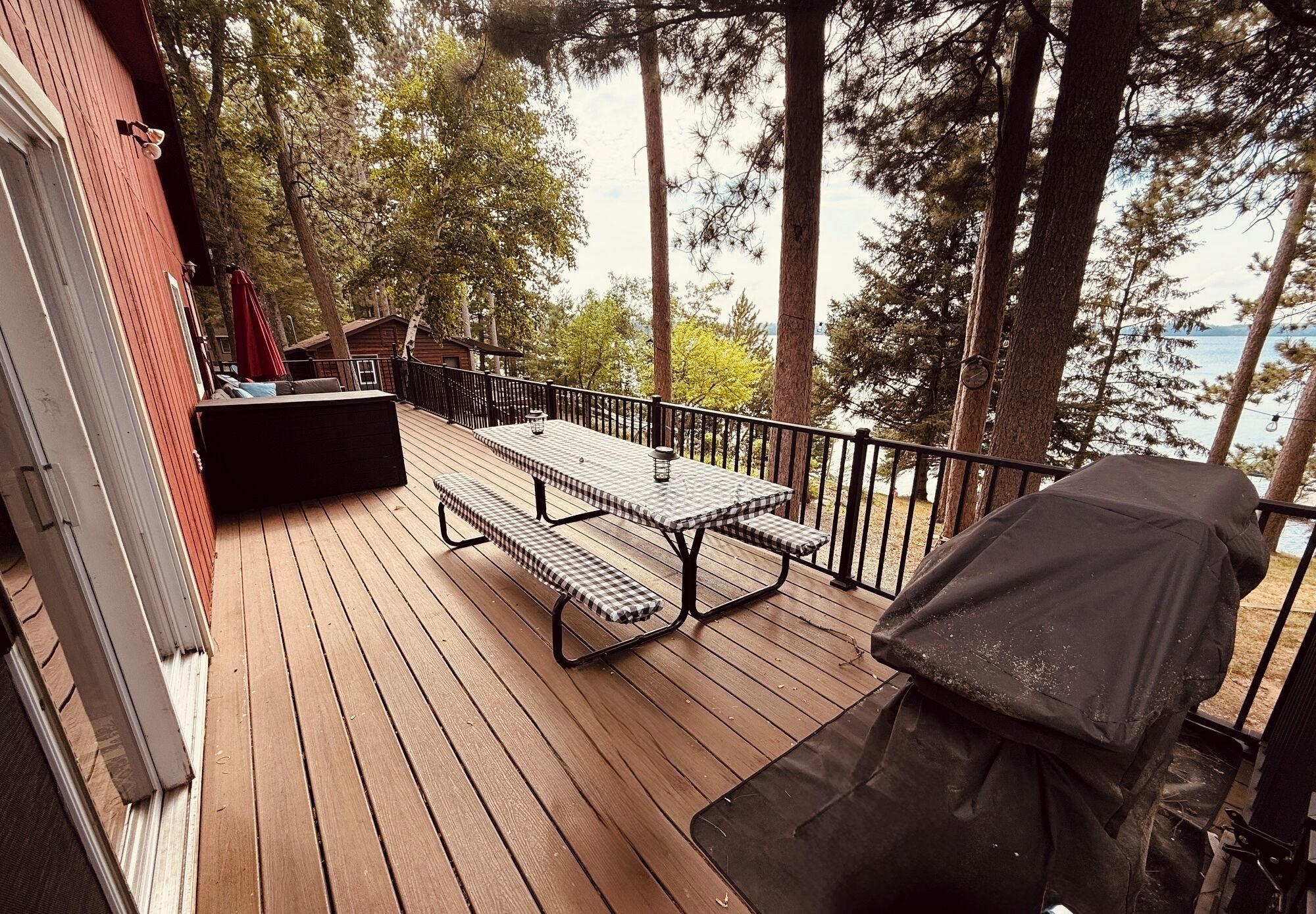 A wooden deck with a picnic table and grill, overlooking a body of water and surrounded by tall trees.