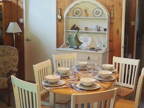 Uma sala de jantar com uma mesa de madeira preparada para seis pessoas, rodeada por cadeiras brancas.