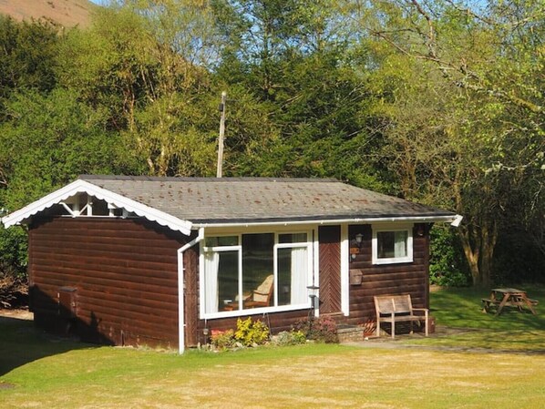 Uma pequena cabana de madeira com telhado em forma de ‘A’, varanda com um banco e uma mesa de piquenique numa área arborizada.