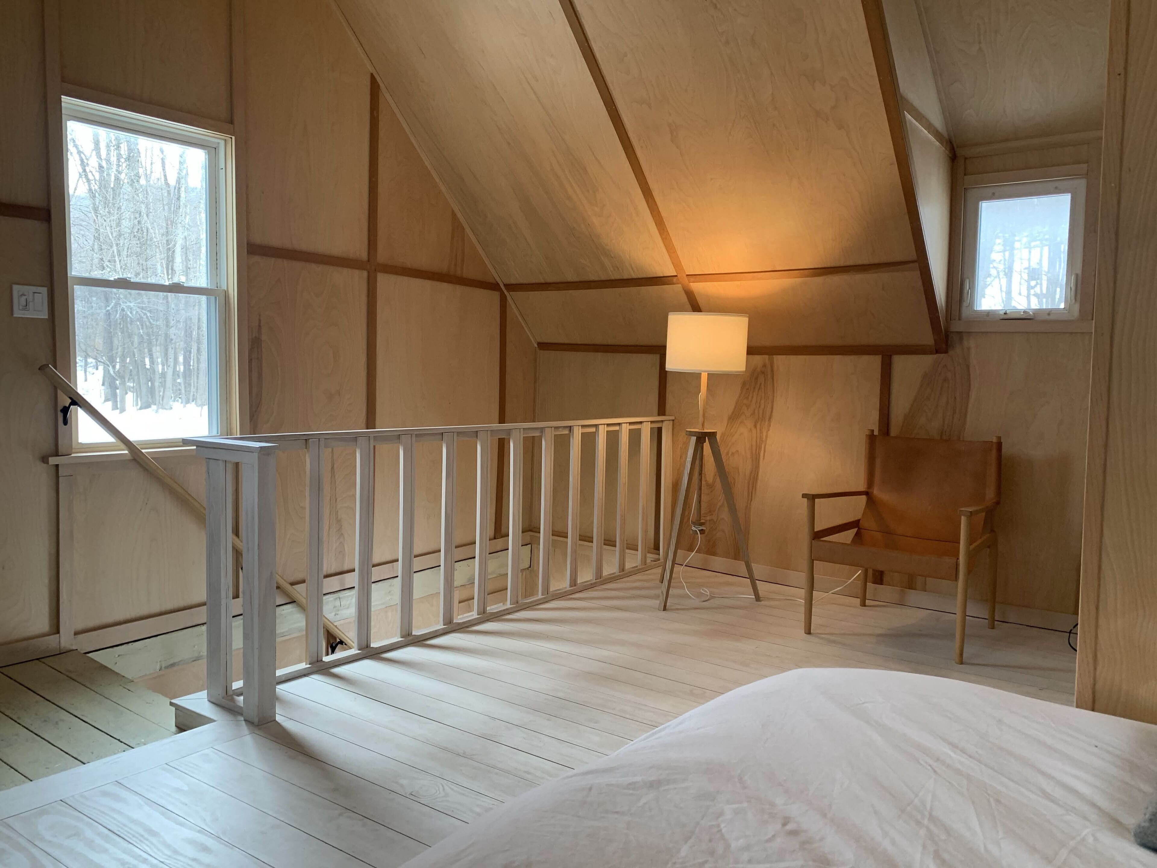 A room with wooden walls and a sloped ceiling, a white bed, a chair, a lamp, and a window with a view of trees.