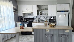 A modern kitchen with white cabinets, a black stove, a microwave, and a refrigerator. There is a small island with stools and a bowl of fruit.
